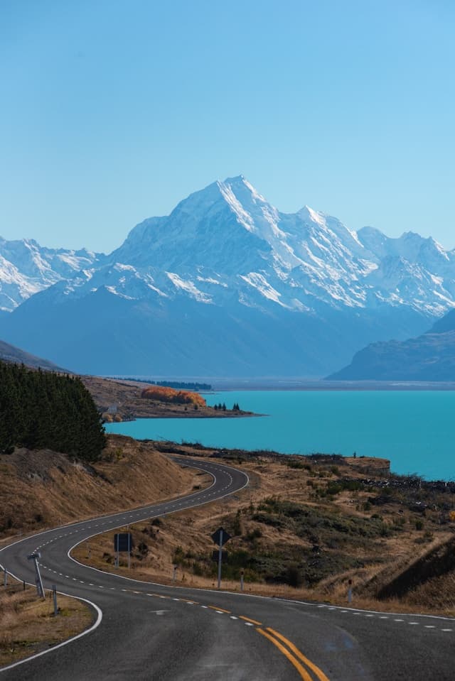 Winding road leading to snow-capped Mount Cook beside turquoise Lake Pukaki, showcasing New Zealand's pristine natural beauty